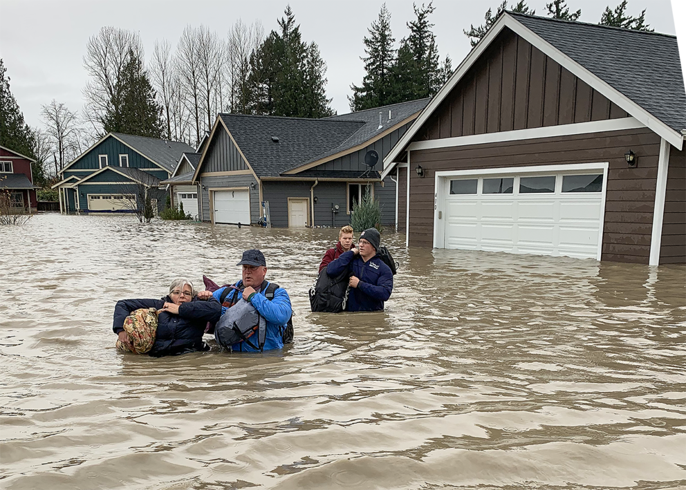 Four people carrying multiple bags cross a flooded street while waist-deep in water