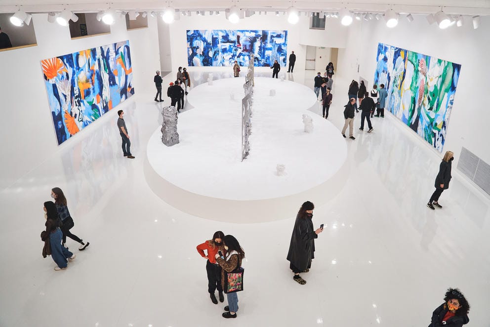 Large exhibition space with a white base in the middle, featuring a metal sculpture. On the three walls are large, colorful paintings