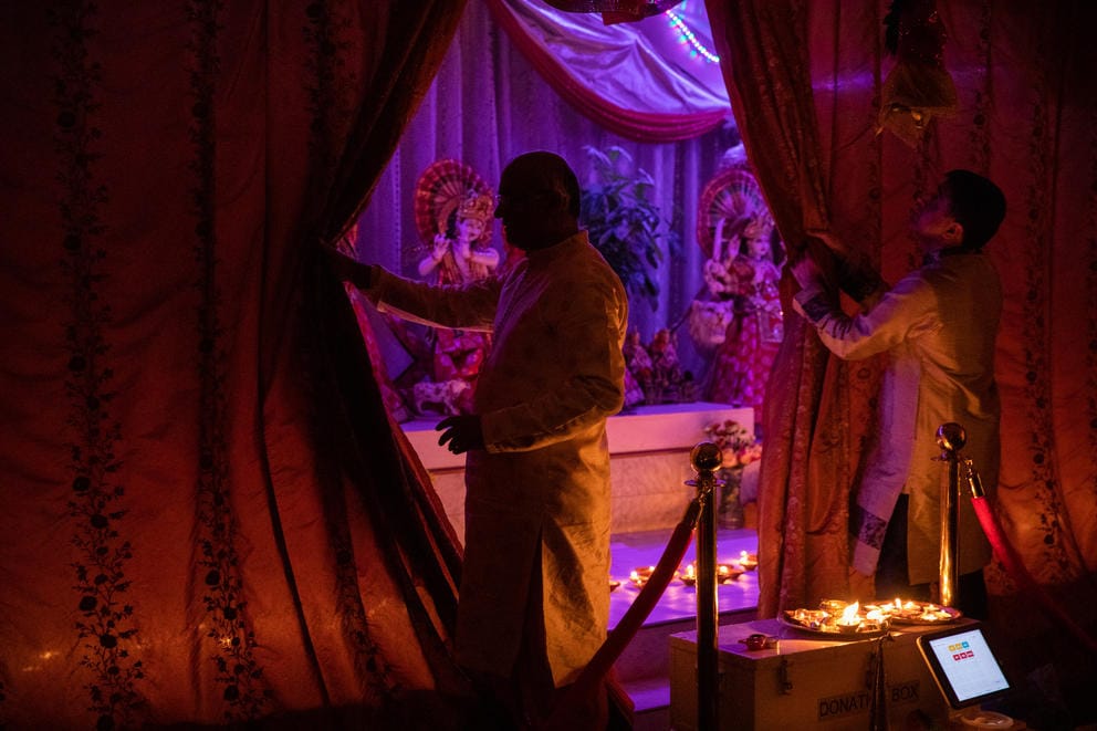 Two men in shadow, open a curtain to reveal a blue-lit stage with deities on it.