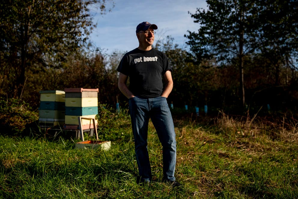 A man stands in an orchard with a beehive behind him