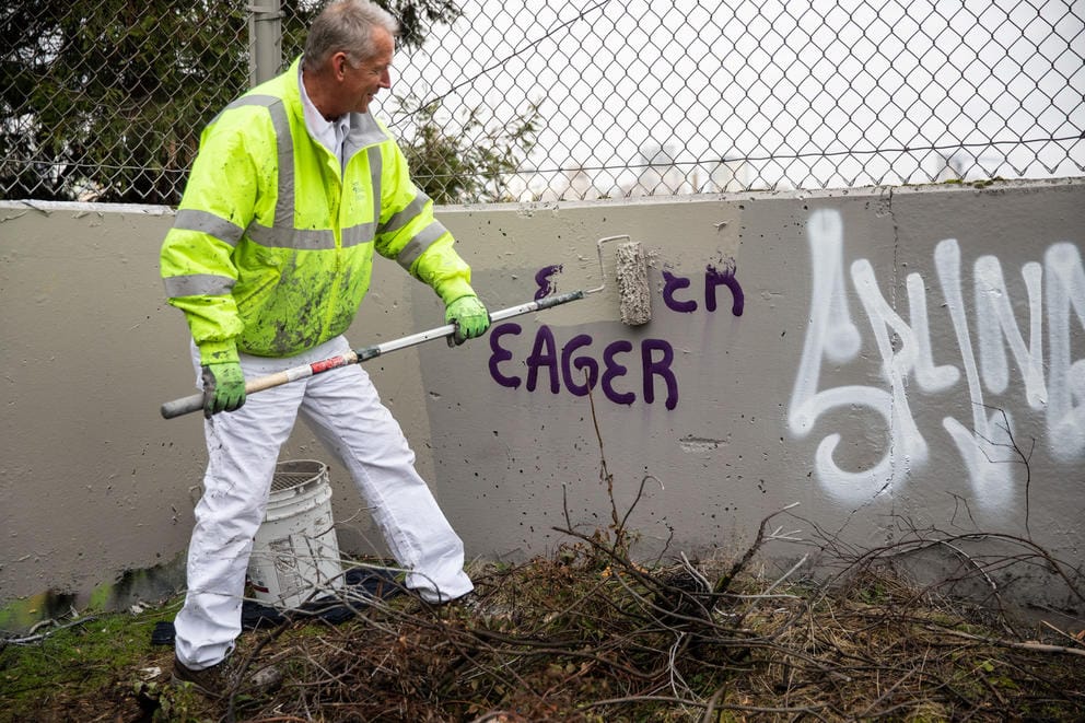 A man in white pants and a hi-vis vest paints over graffiti reading