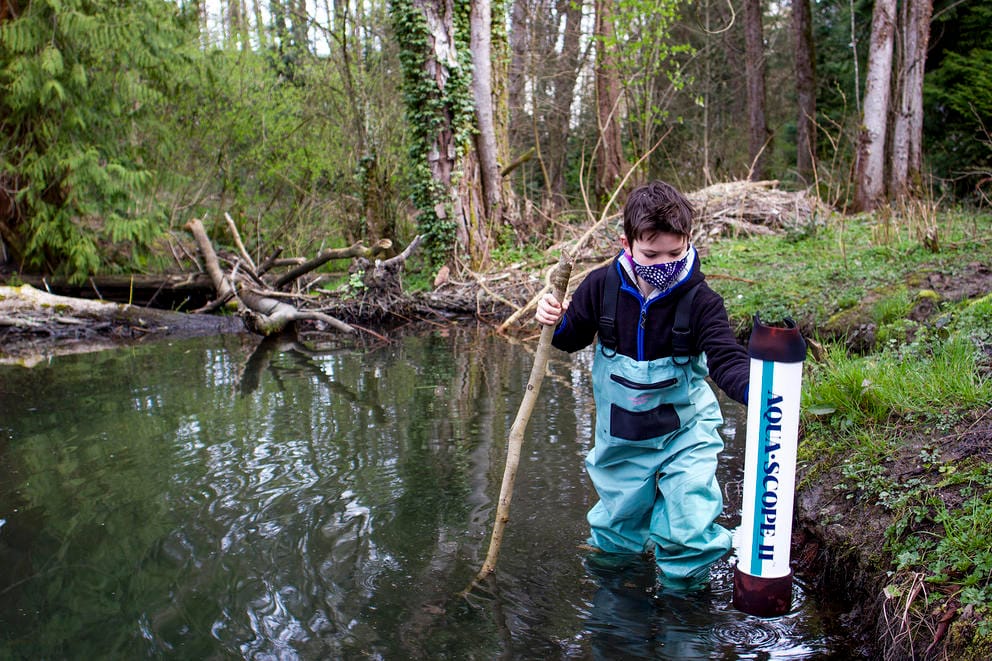 a young boy wearing waders and holding a walking stick and aquascope wades through a pond