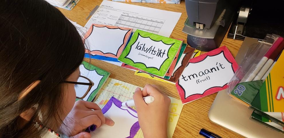 Overhead of a woman writing words — lálu/itsíkt (cream/sugar) and tmaanít (fruit) — on colorful flashcards.