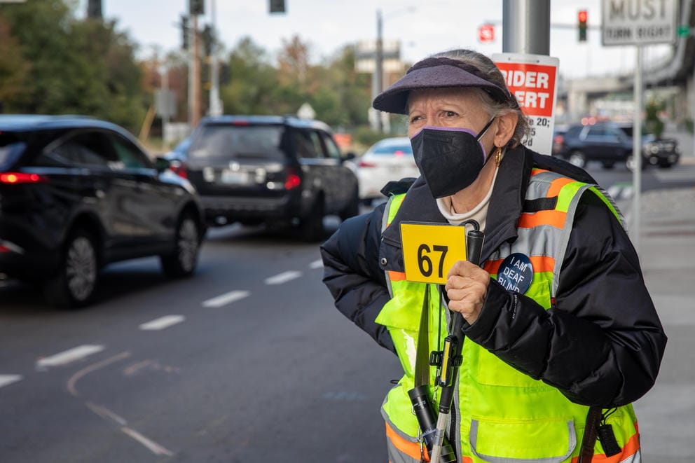A close-up of Lynn Chase, wearing a neon safety vest and black visor, waiting at a bus stop holding a bright yellow card with the number 915 printed on it.