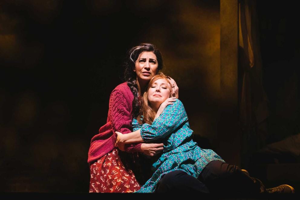 on stage, two women in costume embrace and look fearfully offstage