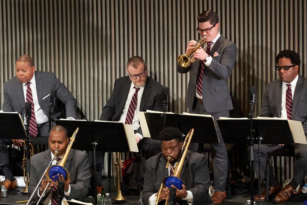 A man in a suit stands and plays the trumpet while other musicians sit in the bandstand