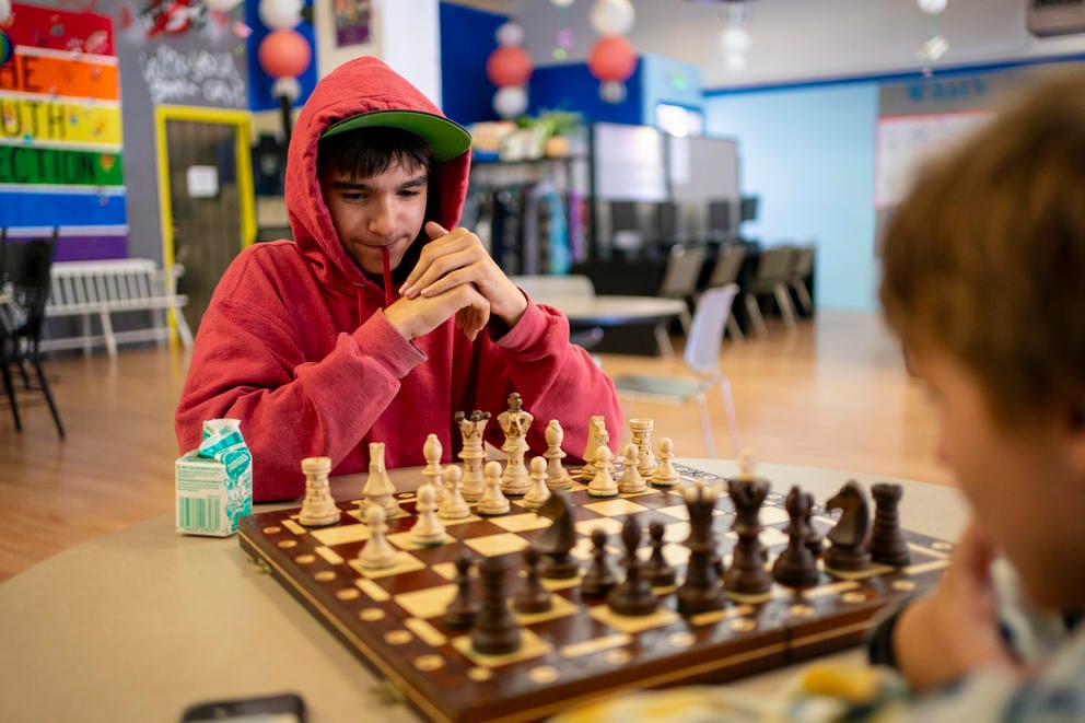 Riley, in a red hoodie sweatshirt, sits looking at a chess board across from another player.