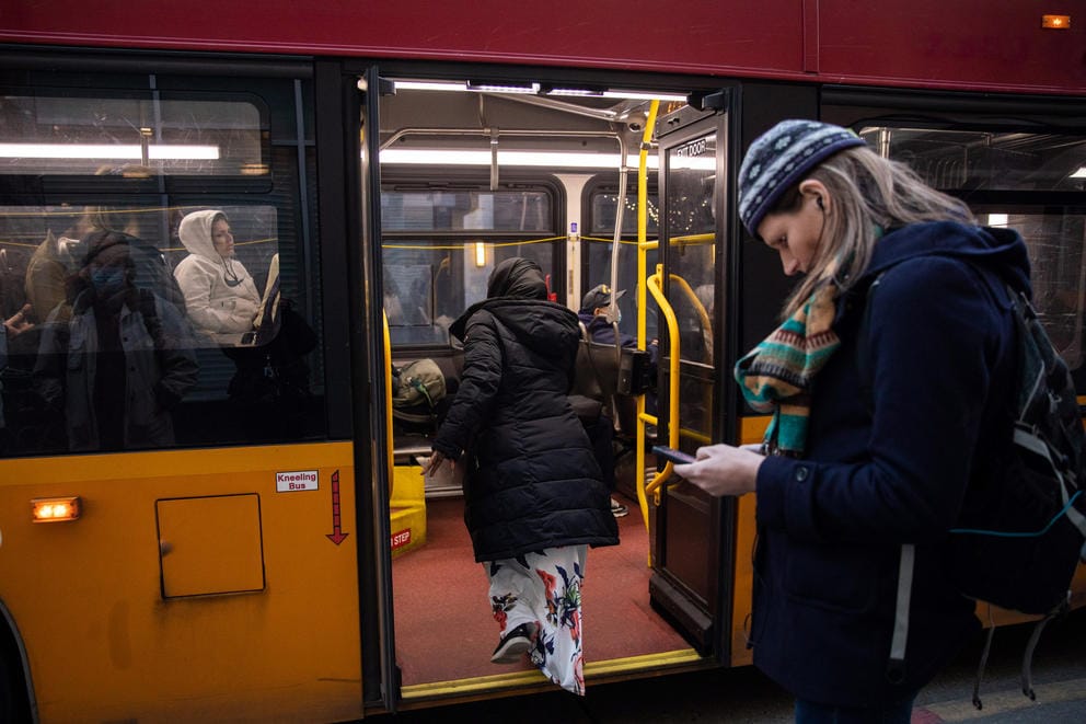 The picture shows two people boarding a red and yellow bus full of people through the back entrance.