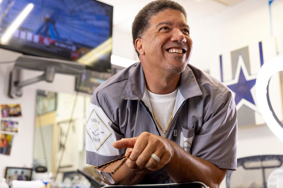 A barber leans forward on his elbows and smiles in his shop.