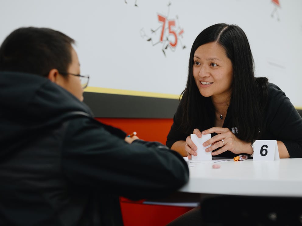 A woman sits at a table speaking to a student sitting across from her.