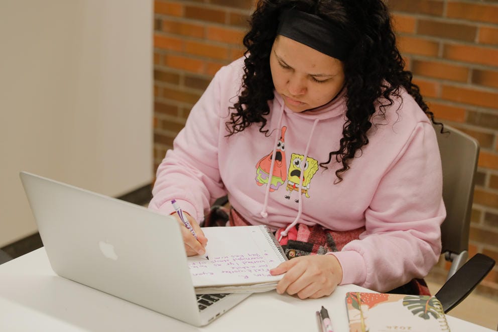 Janell Braxton writes in a notebook that is resting on the keyboard her open laptop.