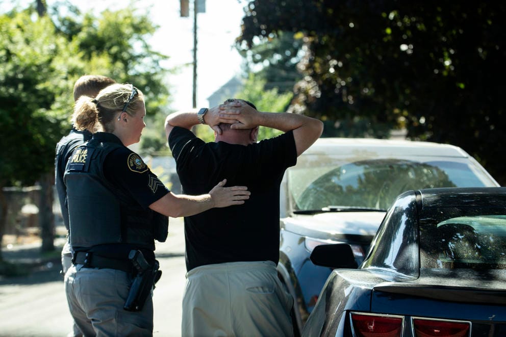 A police officer has a hand on the back of a person who is standing with their hands on their head.