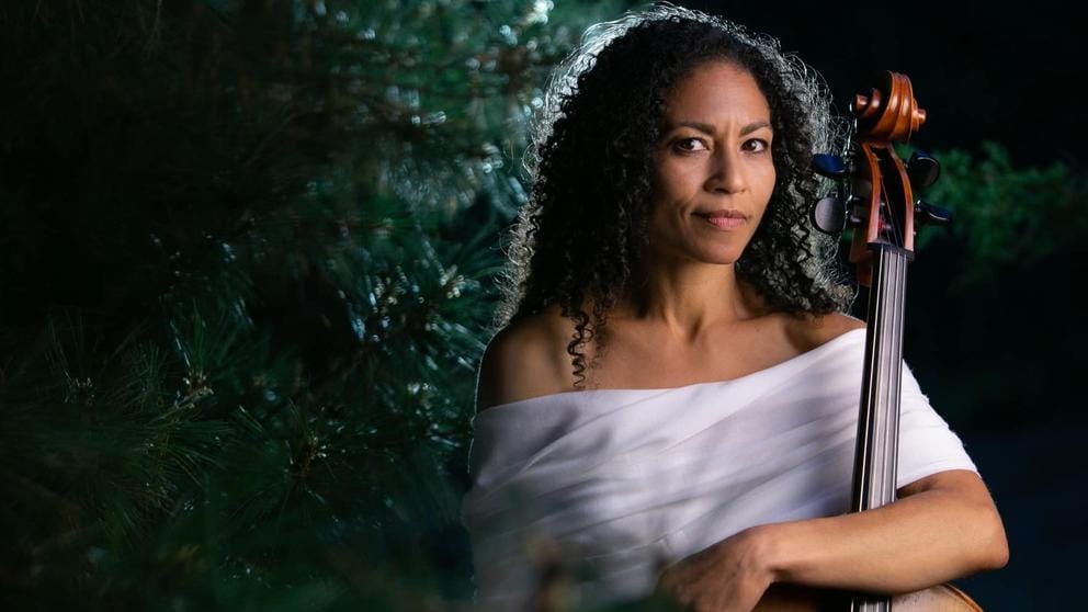 photo portrait of a woman with curly brown hair wearing a white top and holding a cello