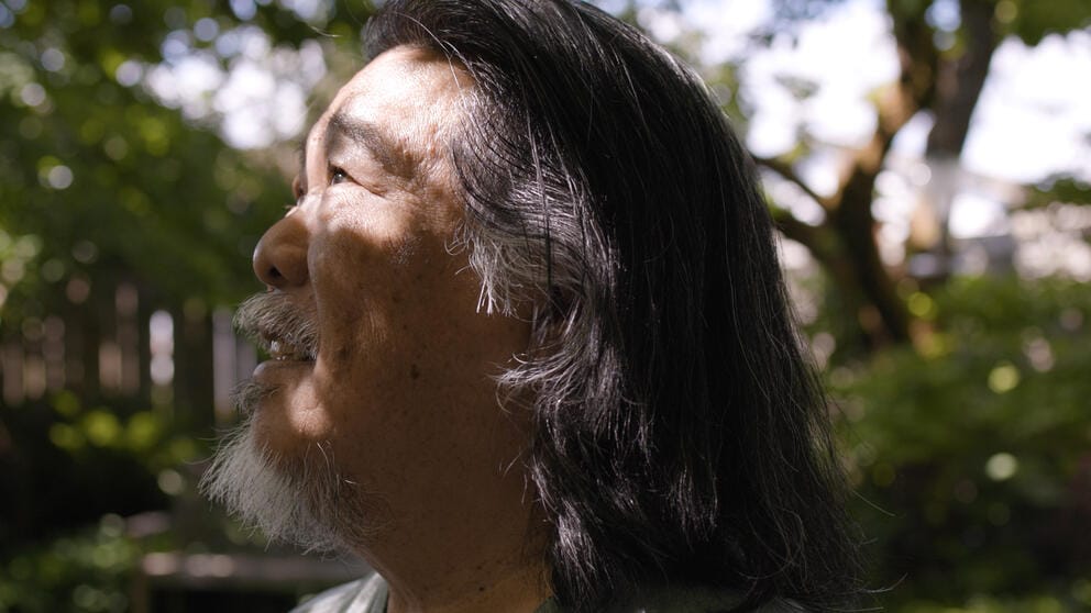 close up of a man with long hair and a beard looking up at dappled light in a garden