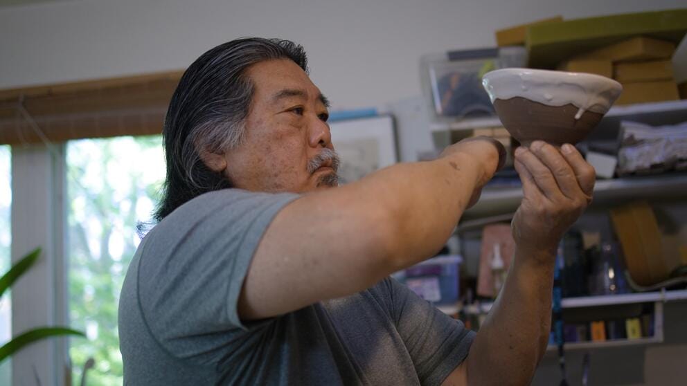 a man with shoulder length black and gray hair holds up a pottery bowl, in the process of glazing it